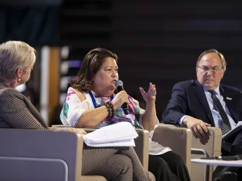 Carmen Auste, CEO of Cancer Warriors Philippines, speaking at a conference panel with two men on either side of her