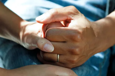 Close up of two hands clasped, resting on a hospital bed, one with a wedding ring