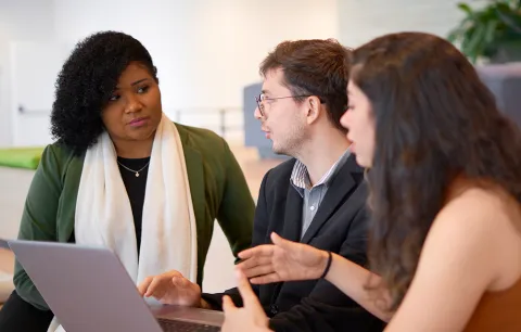 A young white man between a young white woman with long hair on his left and a young black woman with a white scarf on his right, talking in front of a computer