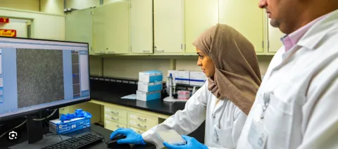 Middle-eastern woman with headscarf and lab coat checking a screen, with a man in lab coat standing just behind her