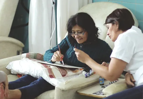 Woman with dark hair and glasses, smiling, is attended by a healthworker as she attempts to draw, while receiving IV treatment and a foot massage