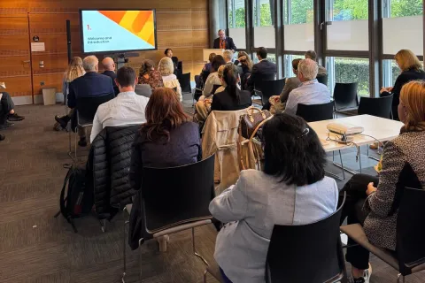 Cary Adams, CEO of UICC presenting at the ATOM Coalition side event. A conference room filled with attendees seated in rows, facing a speaker at the front. The room features large windows allowing natural light, and a presentation screen displaying a colorful slide. The audience appears engaged, with a mix of individuals in business attire, some taking notes or looking at their devices. The setting has a modern design with wooden paneling and a professional atmosphere.