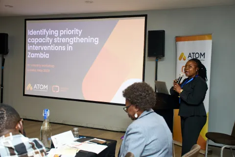 A woman stands at a podium presenting during a workshop titled "Identifying priority capacity strengthening interventions in Zambia." The presentation slide is visible behind her, featuring the workshop title and the ATOM logo. Attendees are seated at tables, engaged in the session, with some taking notes. The setting appears to be a conference room with a professional atmosphere.