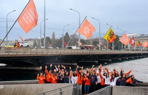 Diverse group of people in branded World Cancer Day sweatshirts standing before the Mont Blanc bridge in Geneva flying World Cancer Day flags