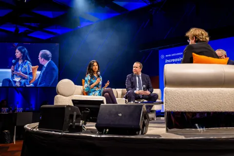 Middle-aged Asian woman and Caucasian man speaking at a conference, sitting on a couch facing the moderator