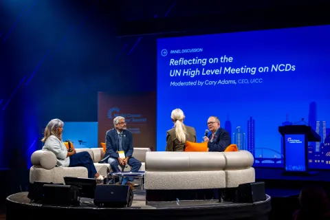 A panel discussion taking place on stage at a conference, featuring four speakers seated in a semi-circle. The backdrop displays the title "Reflecting on the UN High Level Meeting on NCDs," with the moderator, Cary Adams, identified as the CEO of UICC. The stage is illuminated with blue lighting, and a podium is visible on the right. The audience is not shown.