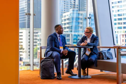 Two men are seated at a small table in a modern, brightly lit space with large windows showcasing a cityscape. One man, dressed in a dark suit and tie, is engaged in conversation with the other, who is wearing a light-colored suit. Both appear to be professionals, and they have bags beside them. They are attendees at the 2025 World Cancer Leaders' Summit in Melbourne, Australia.