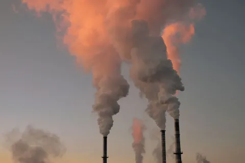 The image shows three tall smokestacks emitting thick plumes of smoke into the sky. The smoke is a mix of white and gray, with hints of pink from the setting sun reflecting off the clouds. The background features a gradient sky transitioning from blue to orange as daylight fades.