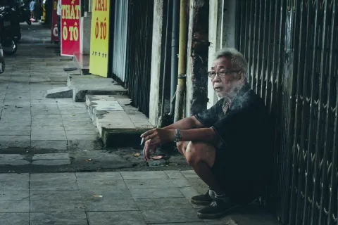 Older man kneeling near a shop, smoking a cigarette
