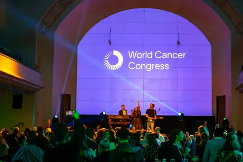Band sings in front of a crowd, backlit by a purple screen with the words "World Cancer Congress". Social event at the World Cancer Congress