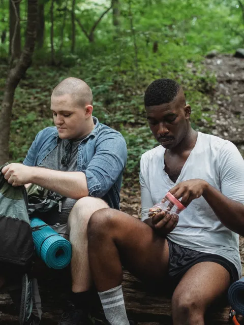 A White and a Black man taking a break to eat a healthy snack while on a hike in a forest