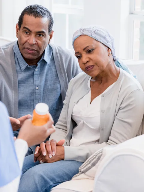 Black male and female couple holding hands, the woman wearing a headscarf indicating illness, both are listening to a doctor