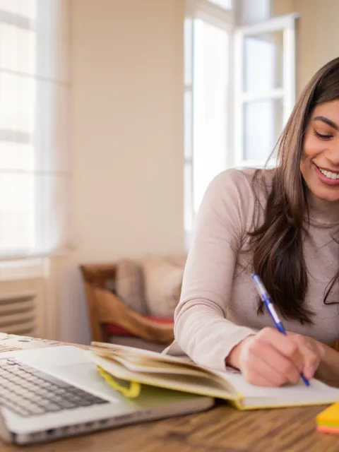 A smiling Hispanic woman writing in a notepad in front of a computer