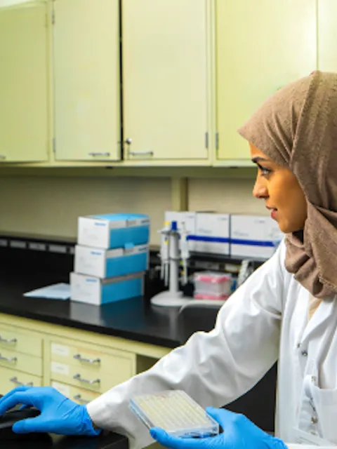 Middle-eastern woman with headscarf and lab coat checking a screen, with a man in lab coat standing just behind her