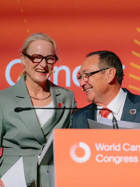 UICC President Ulrika Årehed Kågström wearing a gray suit, smiling with former UICC president Jeff Dunn with a red tie, head turned towards Ulrika, also smiling. both are at the podium at the World Cancer Congress.