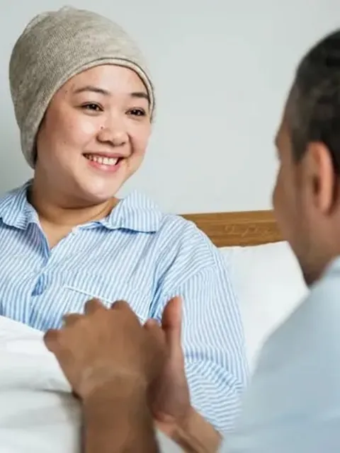 Man seen from behind holding the hand of a smiling woman in a hospital bed, wearing a cap