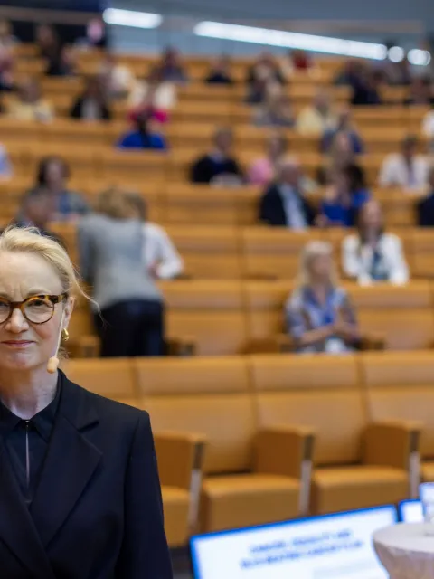 Scandinavian blond woman with glasses standing with her back to an auditorium.