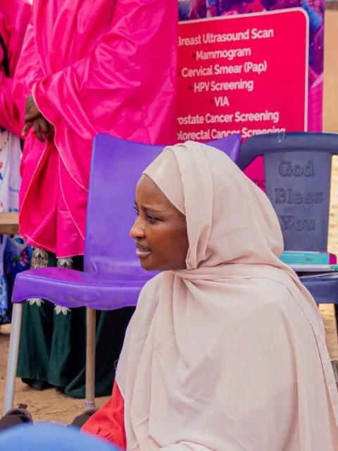 A group of African women wearing bright pink garments, gathered around a sign that lists various medical services, including HPV vaccination for cervical cancer