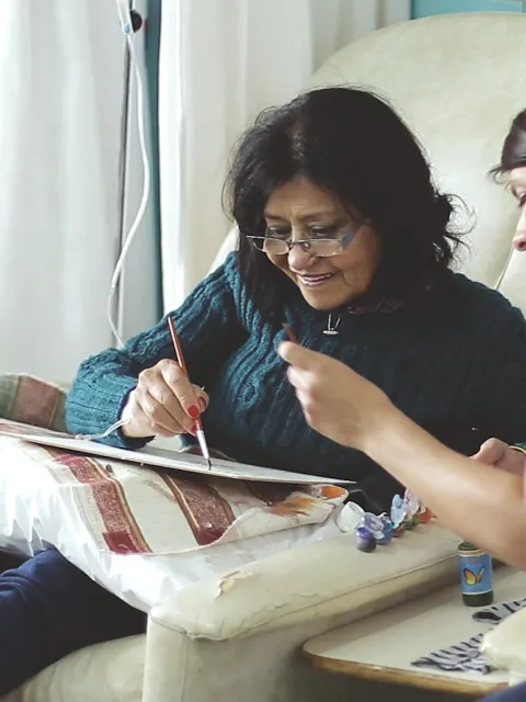 Woman with dark hair and glasses, smiling, is attended by a healthworker as she attempts to draw, while receiving IV treatment and a foot massage