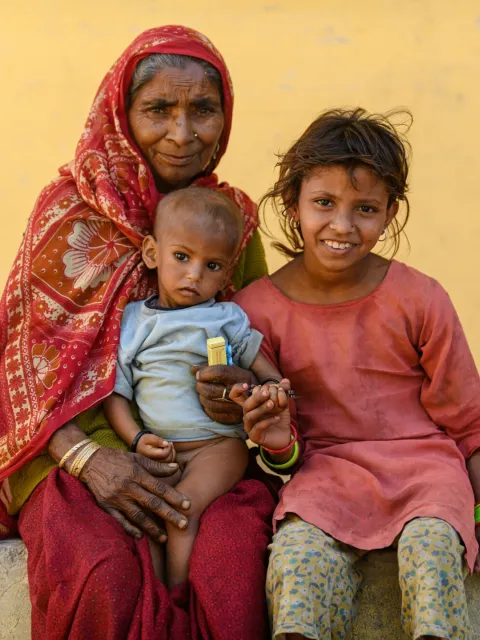 Older Nepalese woman with headdress holding a baby on her knees, a young girl at her side