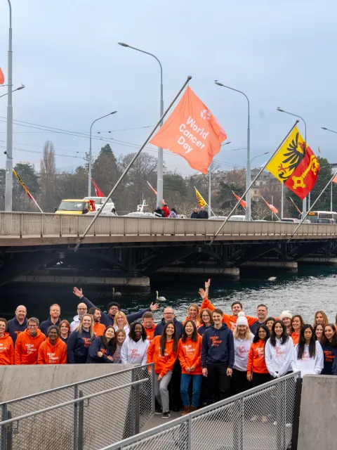 Group of people in World Cancer Day branded white, blue and orange sweatshirts, outside below a bridge flying World Cancer Day flags