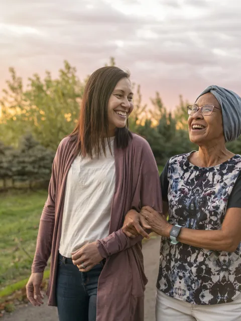 A senior woman with cancer walks with her adult daughter at sunset down a rural road. The affectionate pair are talking and walking with arms linked.