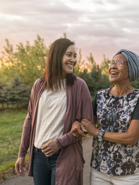 A senior woman with cancer walks with her adult daughter at sunset down a rural road. The affectionate pair are talking and walking with arms linked.