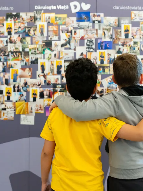 Two children, back to the camera, one in yellow t-shirt, other in great sweatshirt, look at a wall of photos