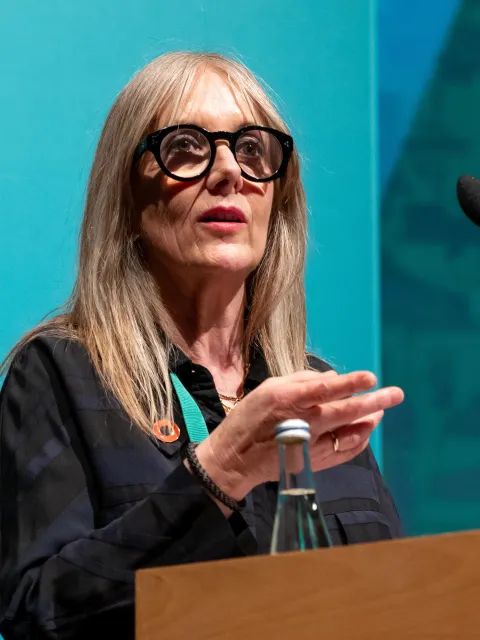 Headshot of a White woman with long blond hair and dark glasses standing at a podium