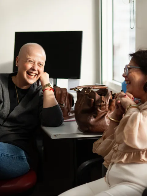 Female cancer patient with no hair laughing as she speaks to another woman on a couch