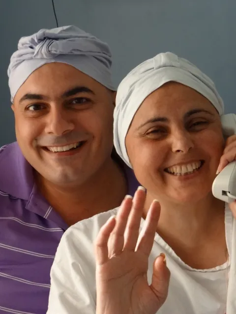Smiling man wearing a purple t-shirt and headscarf with smiling woman with cancer on the phone, a hand raised, also with a headscarf