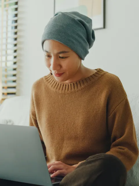 Young Asian woman wearing a headscarf comfortably working on her laptop, seated on a sofa in a well-lit, plant-decorated room.
