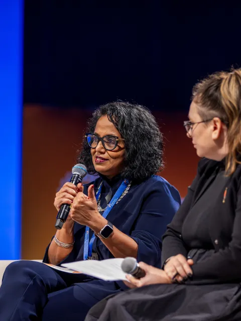 A woman speaking in a mic on a couch, sitting next to another woman