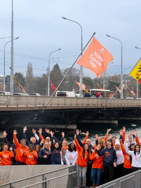 Diverse group of people in branded World Cancer Day sweatshirts standing before the Mont Blanc bridge in Geneva flying World Cancer Day flags