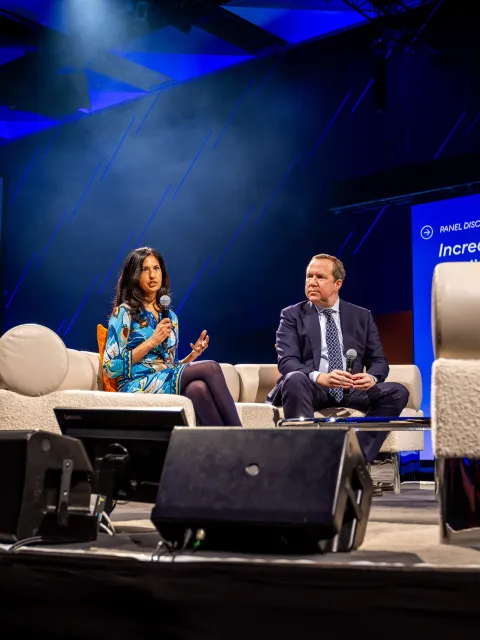 Middle-aged Asian woman and Caucasian man speaking at a conference, sitting on a couch facing the moderator