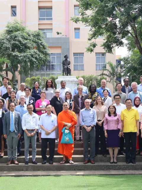 Diverse group of people standing on steps in front of a building surrounded by palm trees, in Bangkok