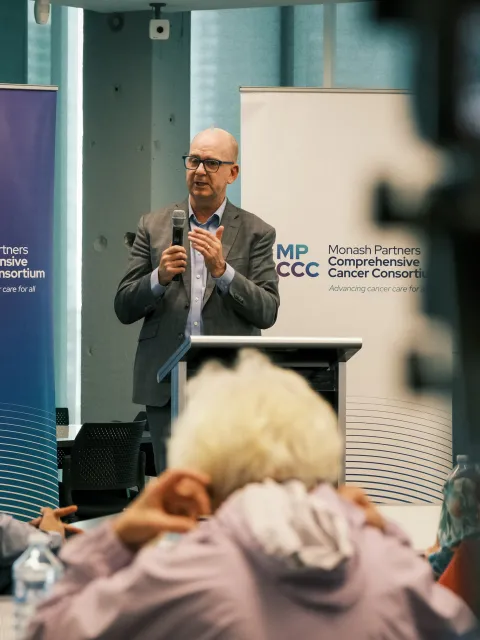 Middle age man wiht no hair, wearing glasses in grey suit with no tie, speaking to a diverse audience