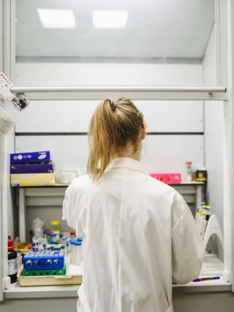Women with long dirty blond hair in a pony tail, in a white lab coat, seen from behind looking at lab equipment