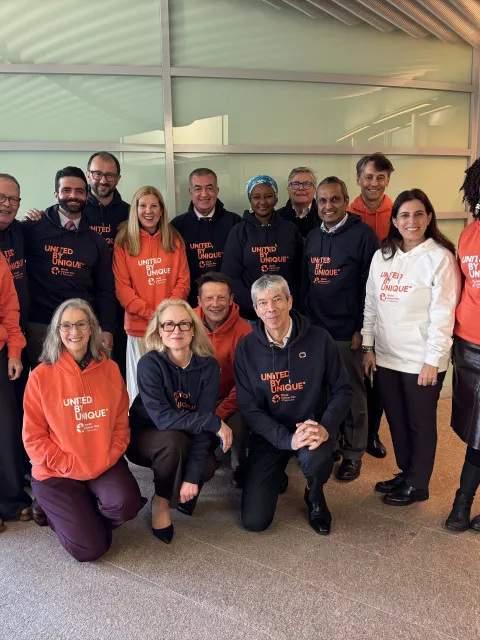 Diverse group of people in two rows, one kneeling in front, posing for the camera in branded white, blue, or orange World Cancer Day United by Unique hoodies