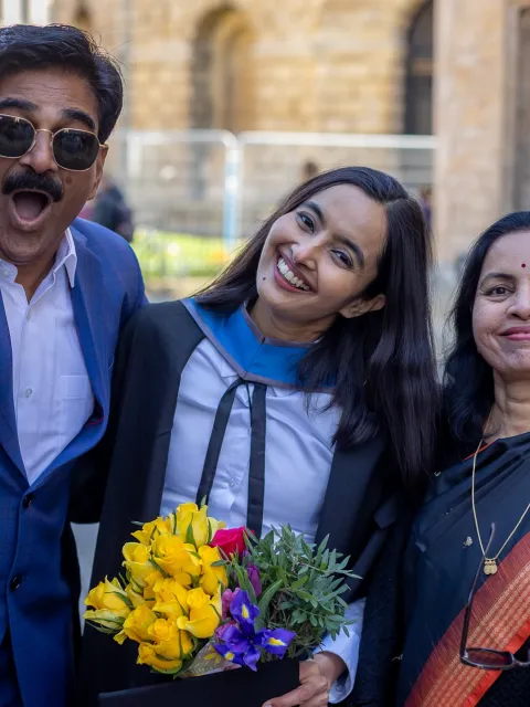 Young Indian woman in graduation gown holding a bouquet of yellow flowers celebrating flanked by her father on her left and mother on her right