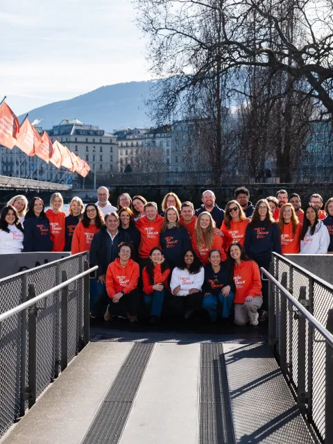 A large group of UICC Staff members, are posing for a photograph on a modern, grey footbridge over a river. They are wearing orange t-shirts with the text "World Cancer Day" printed on them. In the background, a snow-capped mountain range is visible, and several orange flags bearing the "World Cancer Day" logo are displayed along the bridge. The scene is taken at the Mont-Blanc bridge in Geneva, Switzerland