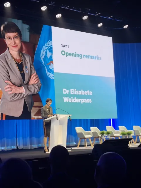 Middle aged white woman at a podium on a big stage with her picture and name Elisabete Weiderpass in the background