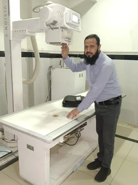 Middle Eastern man in shirt and trousers in front of a radiotherapy machine