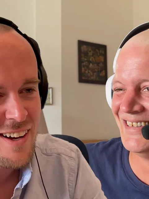 two men wearing headphones with mics, smiling, one without hair, a cancer patient