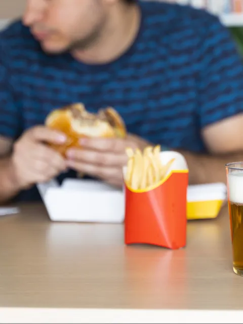 Cropped head of a white male eating burger, fries, soda and beer in front of his computer.