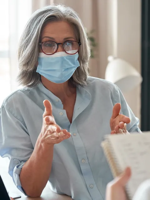 Woman wearing a mask speaking to another person holding a notepad.