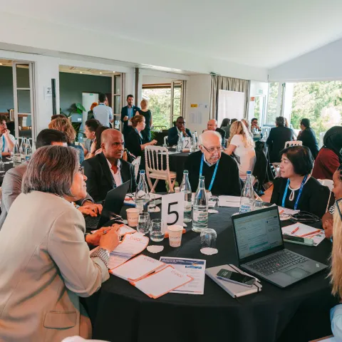 A diverse group of professionals engaged in discussion around a large round table in a well-lit conference room. Participants are seated, with some taking notes and others using laptops. A number "5" is displayed on a stand at the center of the table. In the background, additional attendees are visible, interacting in a collaborative environment. Large windows allow natural light to fill the space, creating an open and inviting atmosphere.