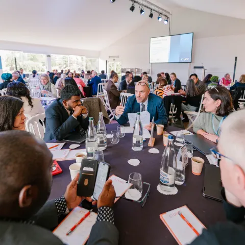 A group of business professionals are gathered around a dark-colored table in a large white tent. Several individuals are taking notes and using smartphones, while one man gestures as he speaks. A large screen displays a presentation, and other attendees are visible in the background, suggesting a conference or workshop setting.