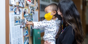 Women with her child both wearing masks in a cancer ward looking at a picture board, with the child pointing at a picture of three doctors