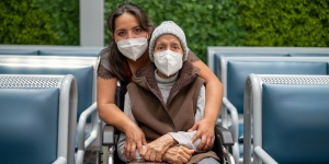 Young woman standing behind and embracing an older woman in a wheelchair, both wearing masks. Photo by Adán Jardón (c) UICC 2021